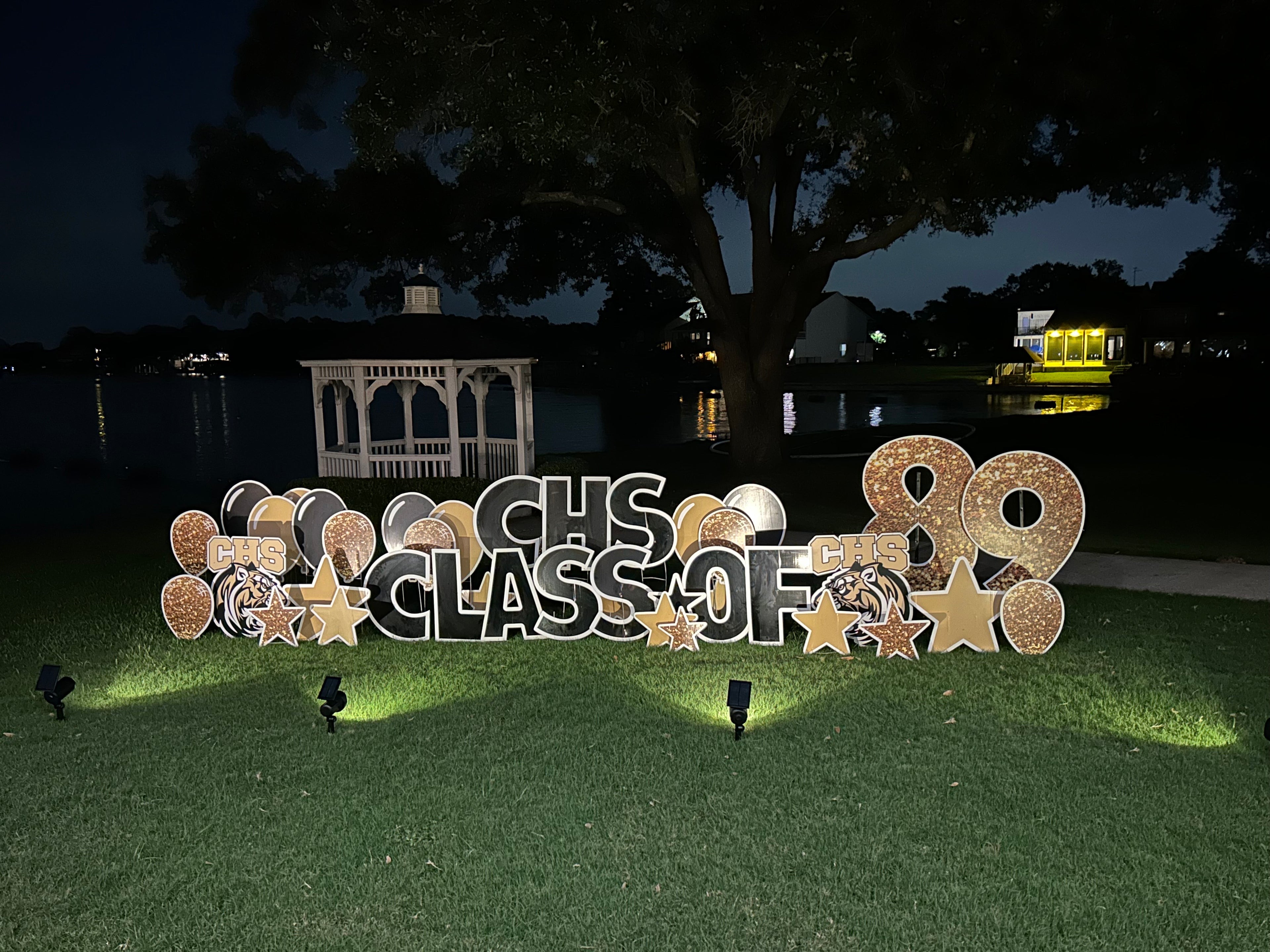 A vibrant yard card display by Texas Party in the Yard, designed for a class reunion event. The setup features a personalized "Class Reunion" message, class year, school colors, and nostalgic graphics like graduation caps and school mascots, night lighting for the sign, creating a welcoming and festive atmosphere for alumni gathering in Montgomery, TX.