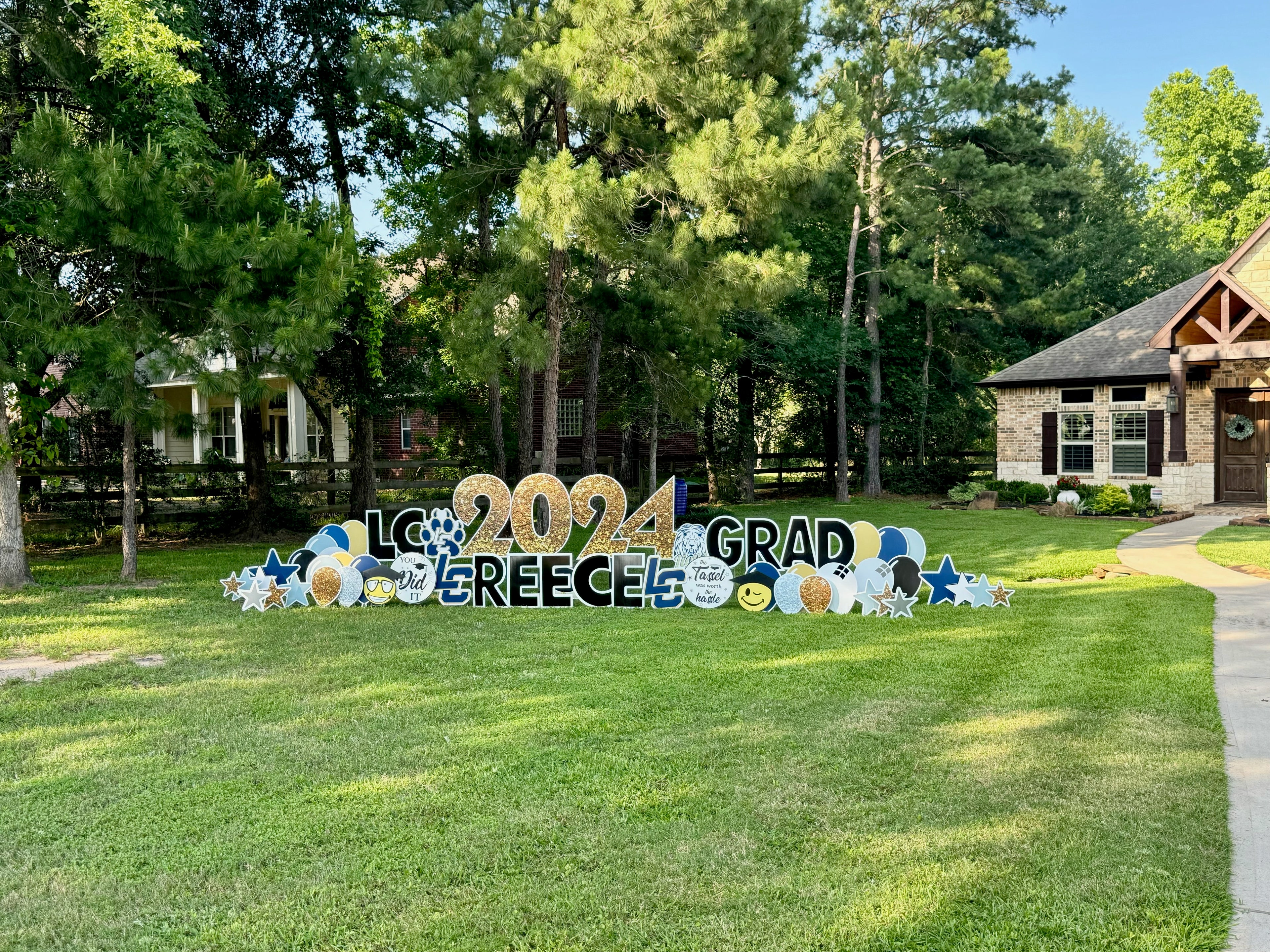 A celebratory yard card setup by Texas Party in the Yard, marking a high school graduation. The display includes a "Congrats Grad" message, the graduate's name, graduation caps, diplomas, and school colors, arranged in a front yard in Montgomery, TX.