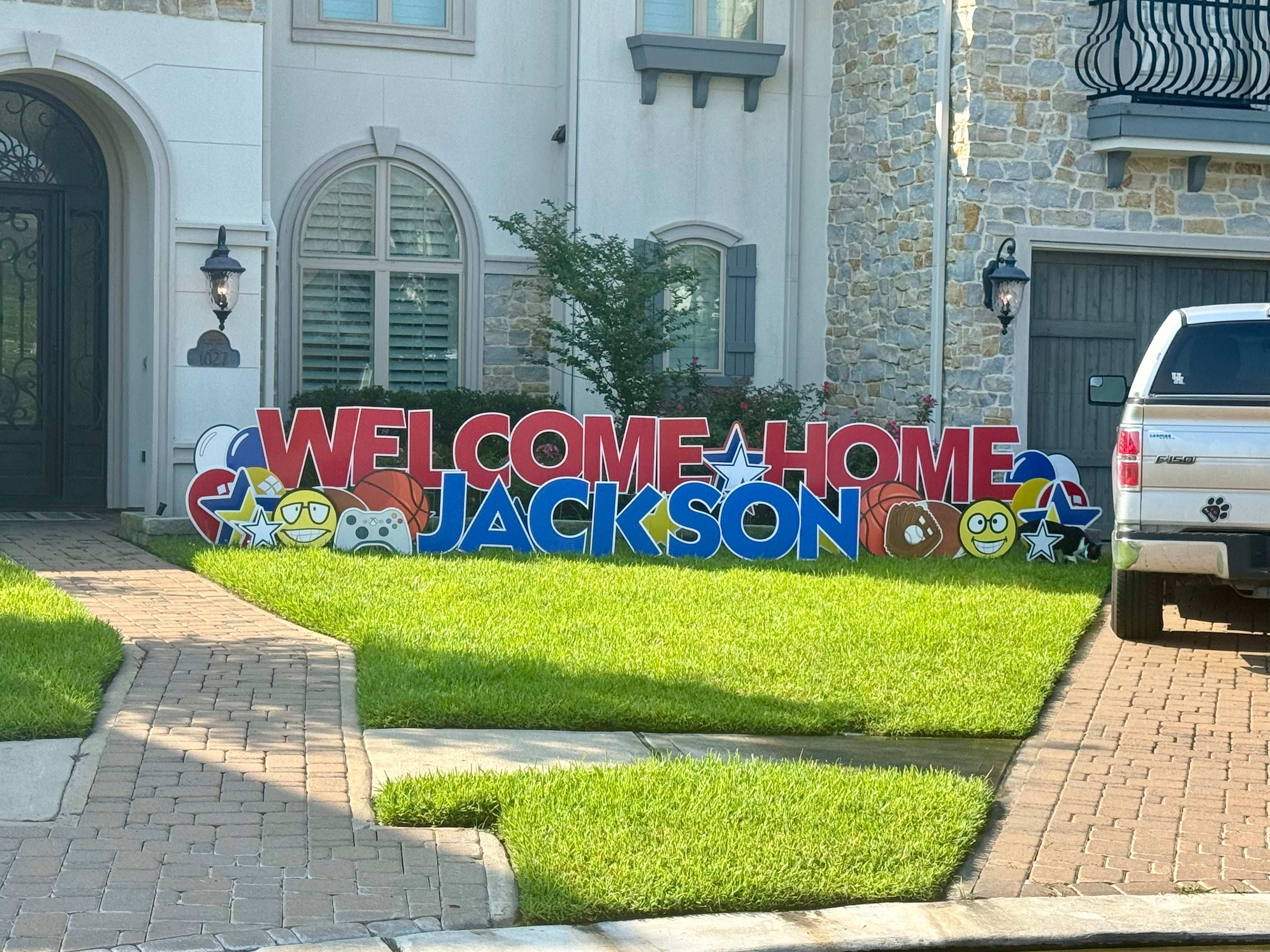 A warm yard card display by Texas Party in the Yard, celebrating a welcome home event. The setup includes a "Welcome Home" message with the person's name, balloons, stars, and heartwarming graphic images, creating a festive atmosphere in Montgomery, TX.