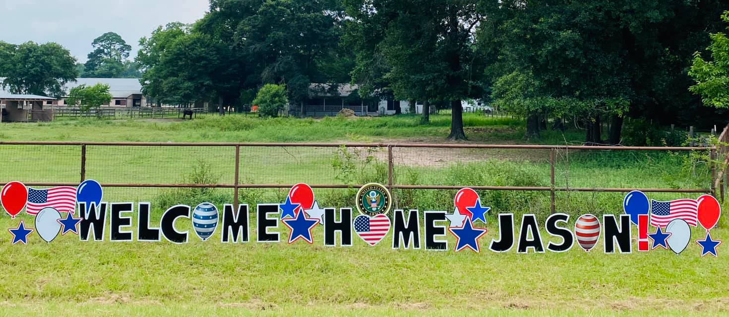 A proud yard card display by Texas Party in the Yard, celebrating the return of a military member. The setup includes a "Welcome Home" message with the service member's name, American flags, stars, and military-themed graphics, arranged in a front yard in Montgomery, TX.